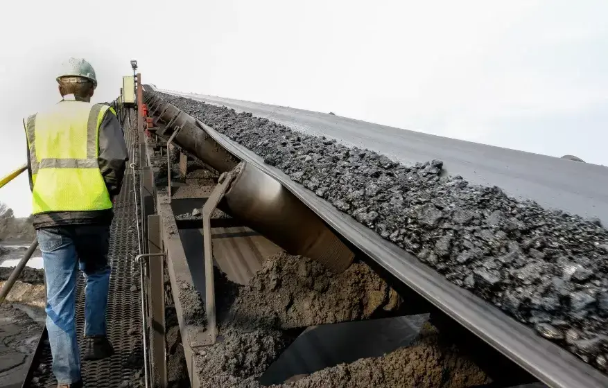 Worker inspecting coal conveyor belt with dust buildup at industrial site