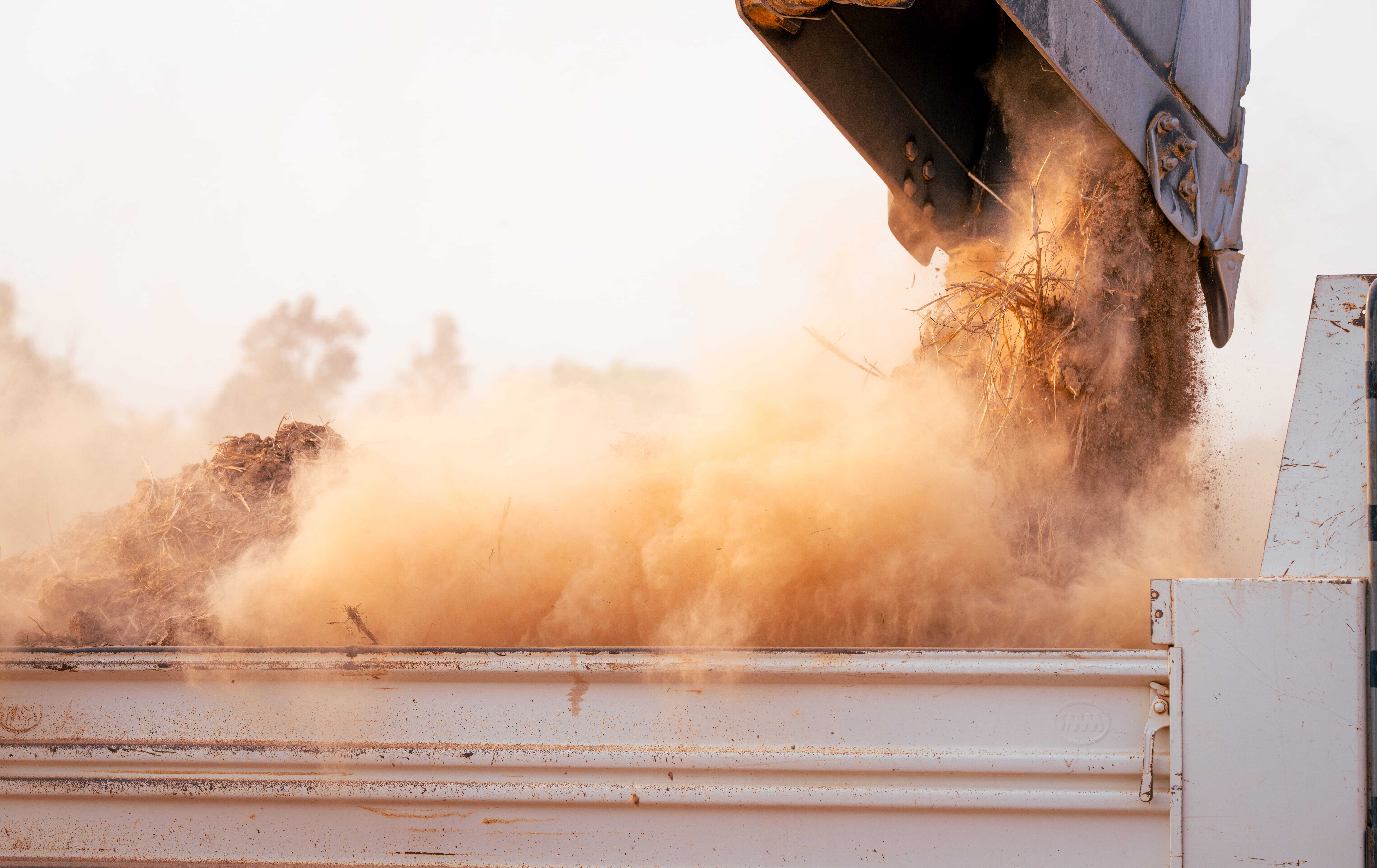 An excavator bucket tipping a dusty material into a truck, creating a large cloud of airborne dust, which can be reduced by using an automatic misting system.