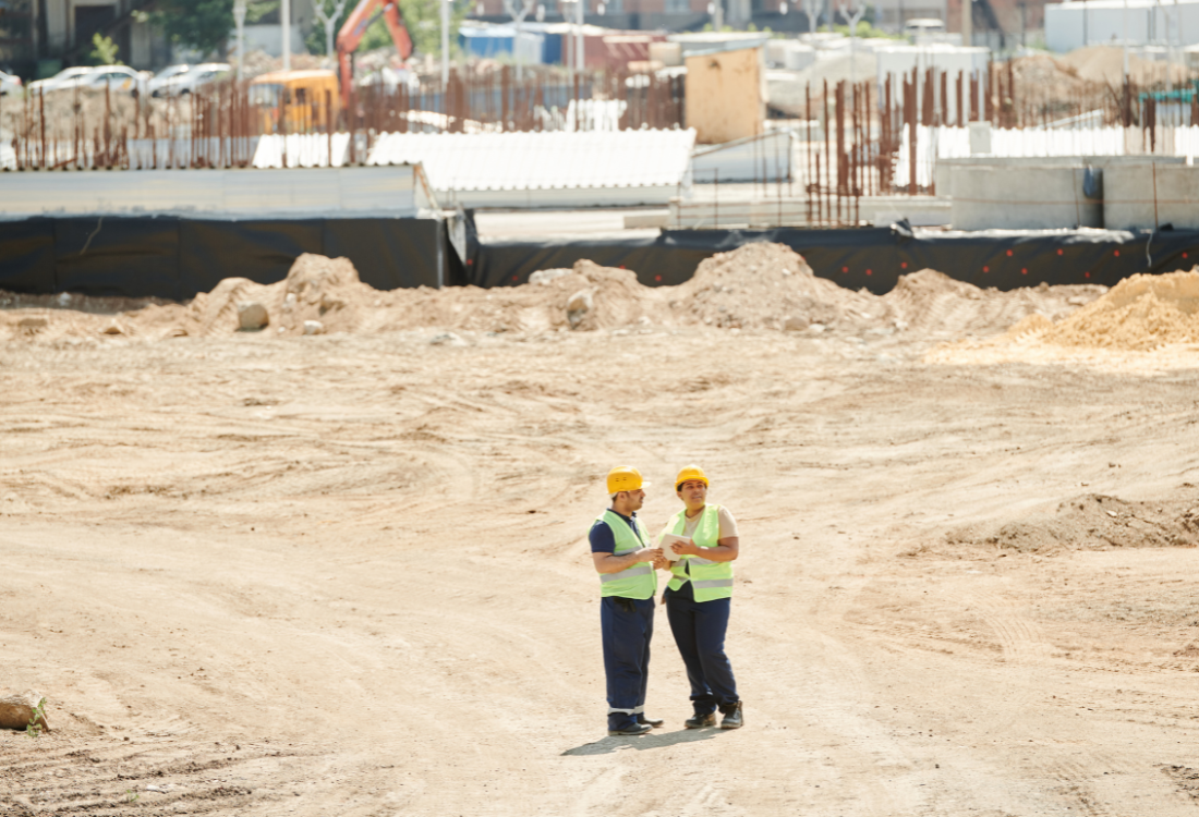 Two industrial safety workers in high visibility vests and hard hats conducting a site survey on a dusty construction site, highlighting the importance of dust suppression systems.