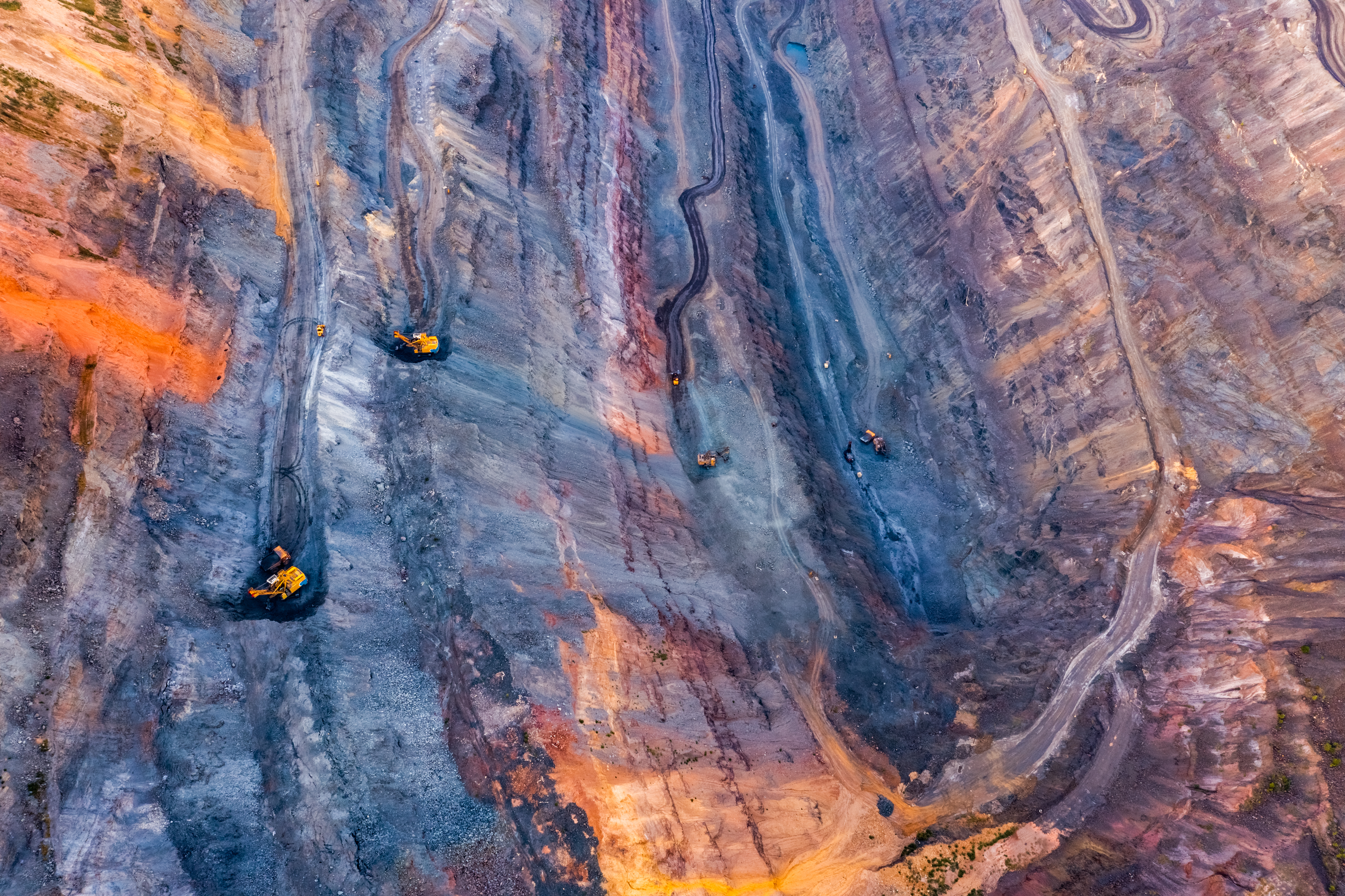 Open pit iron mine with heavy machinery operating across layered ore seams, generating iron ore dust during extraction and crushing operations.